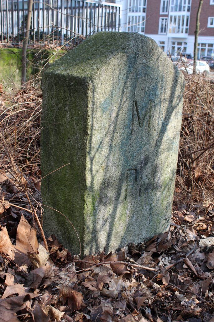 Verwitterter Granit-Grenzstein von 1912 im Laub am Doormannsweg in Hamburg-Eimsbüttel, im Hintergrund Wohnbebauung