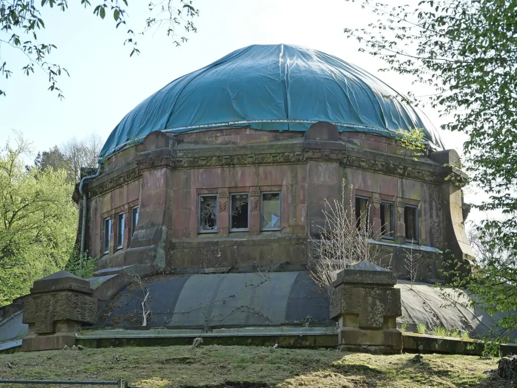 Die Kuppel des Mausoleums unter einer straff gespannten Plane, darunter eine Fensterreihe mit fehlenden Scheiben; auf dem Dachrand wachsen junge Bäume