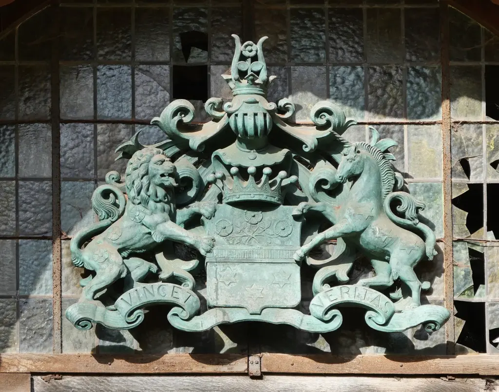 Grün patiniertes Metallwappen über dem Portal, gehalten von Löwe und Einhorn, mit Freiherrnkrone und dem Schriftband VINCET VERITAS; dahinter eine Bleiverglasung mit mehreren fehlenden Scheiben