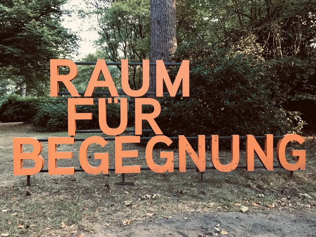 Freistehende orangefarbene Großbuchstaben auf Stahlrohren mit den Worten „Raum für Begegnung“ vor Rhododendren