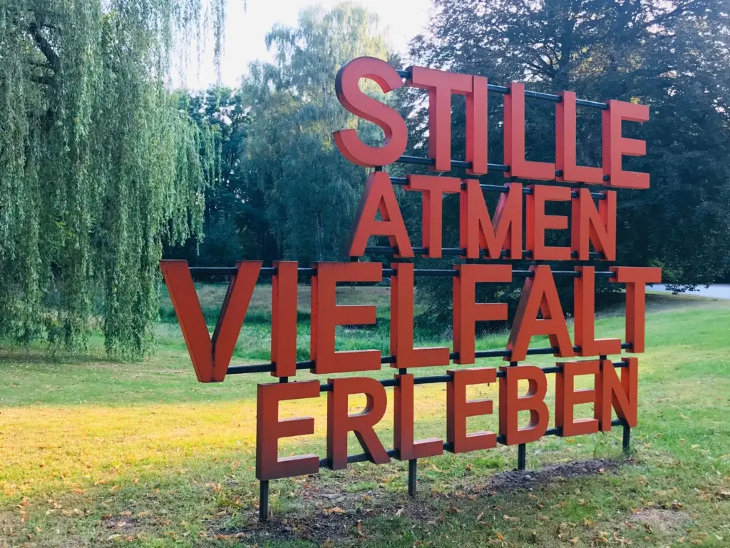 Freistehende orangefarbene Großbuchstaben auf Stahlrohren mit den Worten „Stille atmen“ und „Vielfalt erleben“ auf einer Wiese