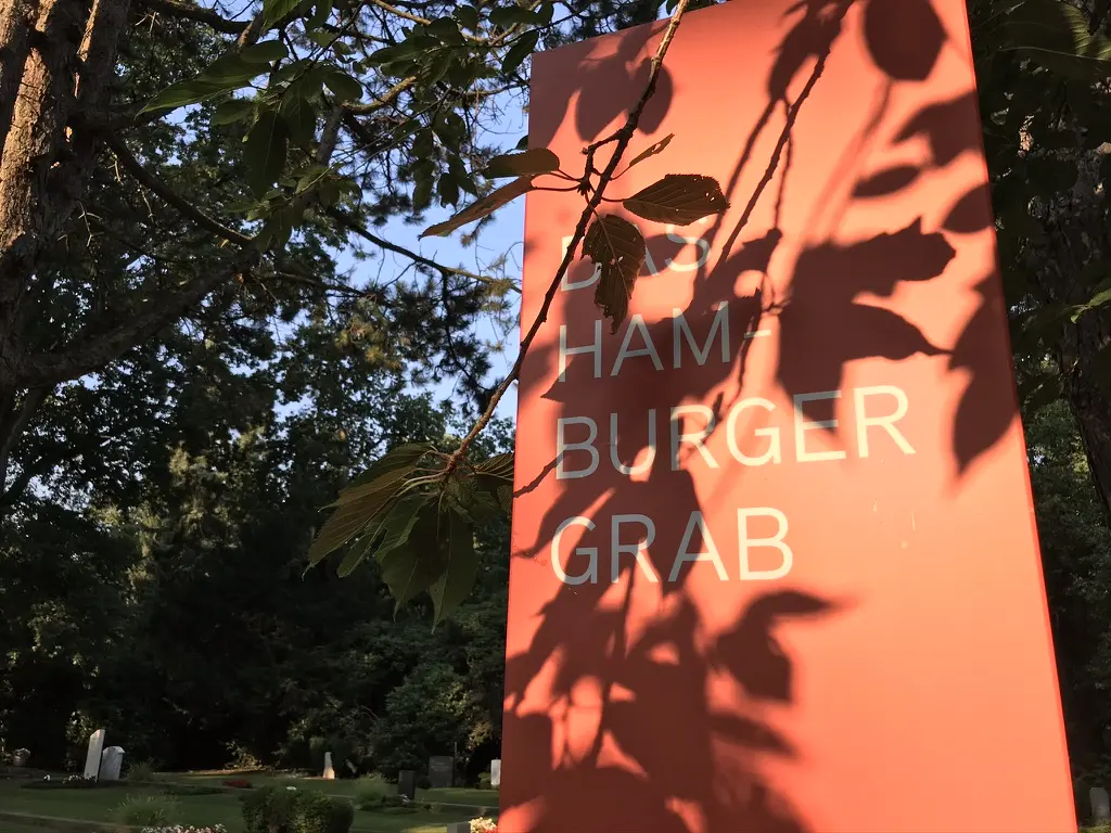 Orangefarbene Stele mit der Aufschrift „Das Hamburger Grab“, mit Blattschatten auf der Fläche, im Hintergrund Grabsteine