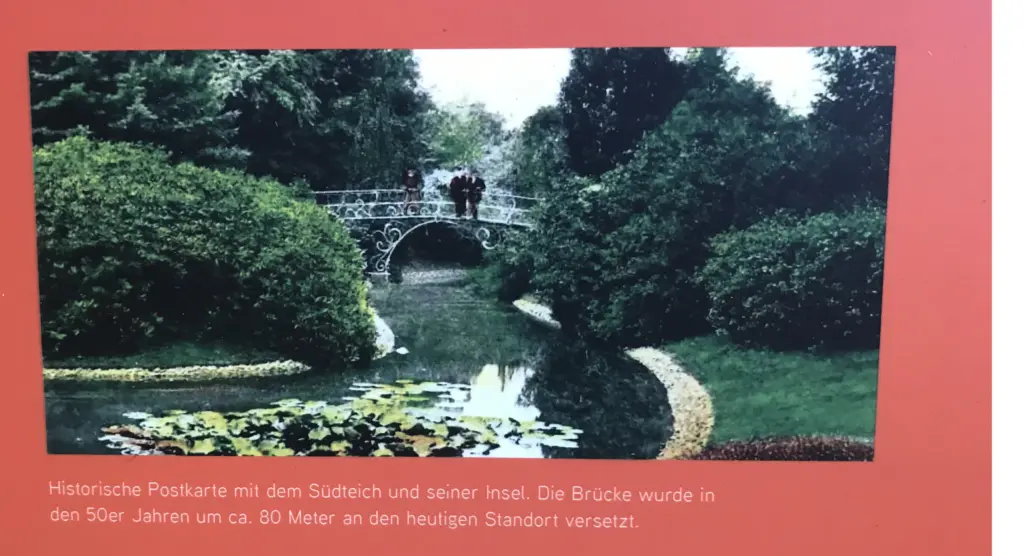 Orangefarbene Informationsstele am Südteich mit einer historischen Postkarte, die den Teich, die Insel und die schmiedeeiserne Brücke mit Personen darauf zeigt