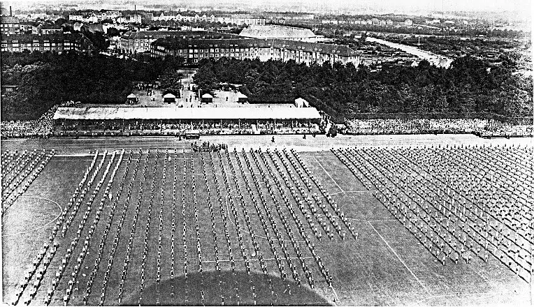 Blick von oben auf ein Sportfeld, auf dem Tausende Schuelerinnen und Schueler in langen Reihen stehen