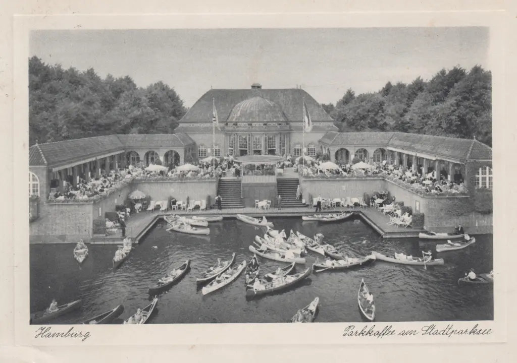 Das Stadtparkcafé vom Stadtparksee aus: Rundbau mit Kuppel zwischen zwei Arkadenflügeln, davor besetzte Terrassen und das Hafenbecken voller Kanus
