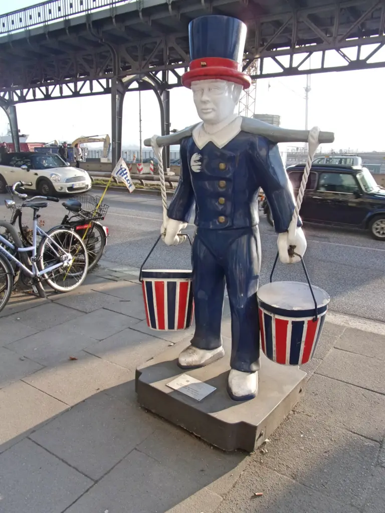 Blau-weiße Wasserträgerfigur mit rot gebändertem Zylinder und rot-blau gestreiften Eimern auf einem Betonsockel am Gehweg des Johannisbollwerks, dahinter die Hochbahnbrücke.